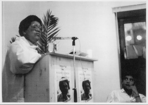 a black and white photograph of Audre Lorde, standing at a podium and giving a lecture