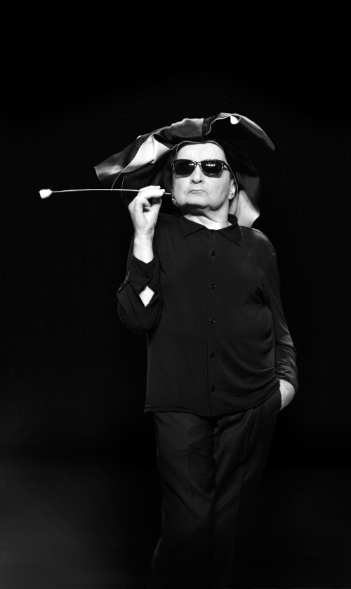 A black and white photograph of the performer Raimund Hoghe standing on a theatre stage